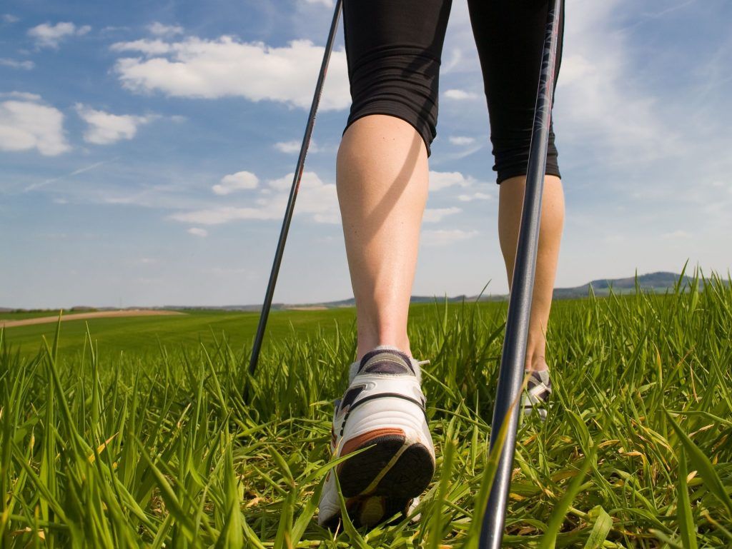 Dettaglio di una persona che pratica Nordic Walking con i bastoncini in un prato sotto il cielo azzurro